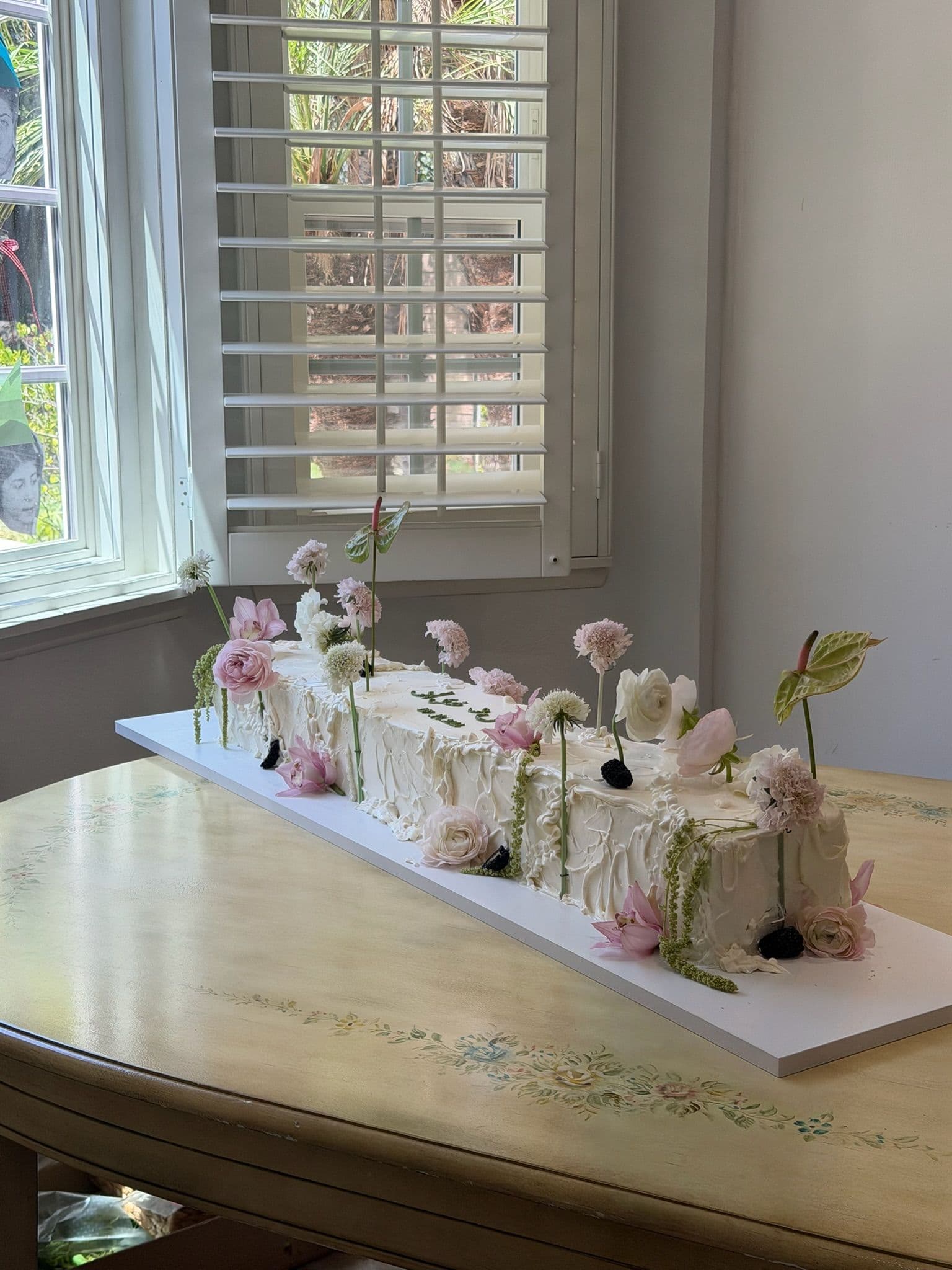 Long floral sheet cake on a painted table, flowers standing on tall stems along its length