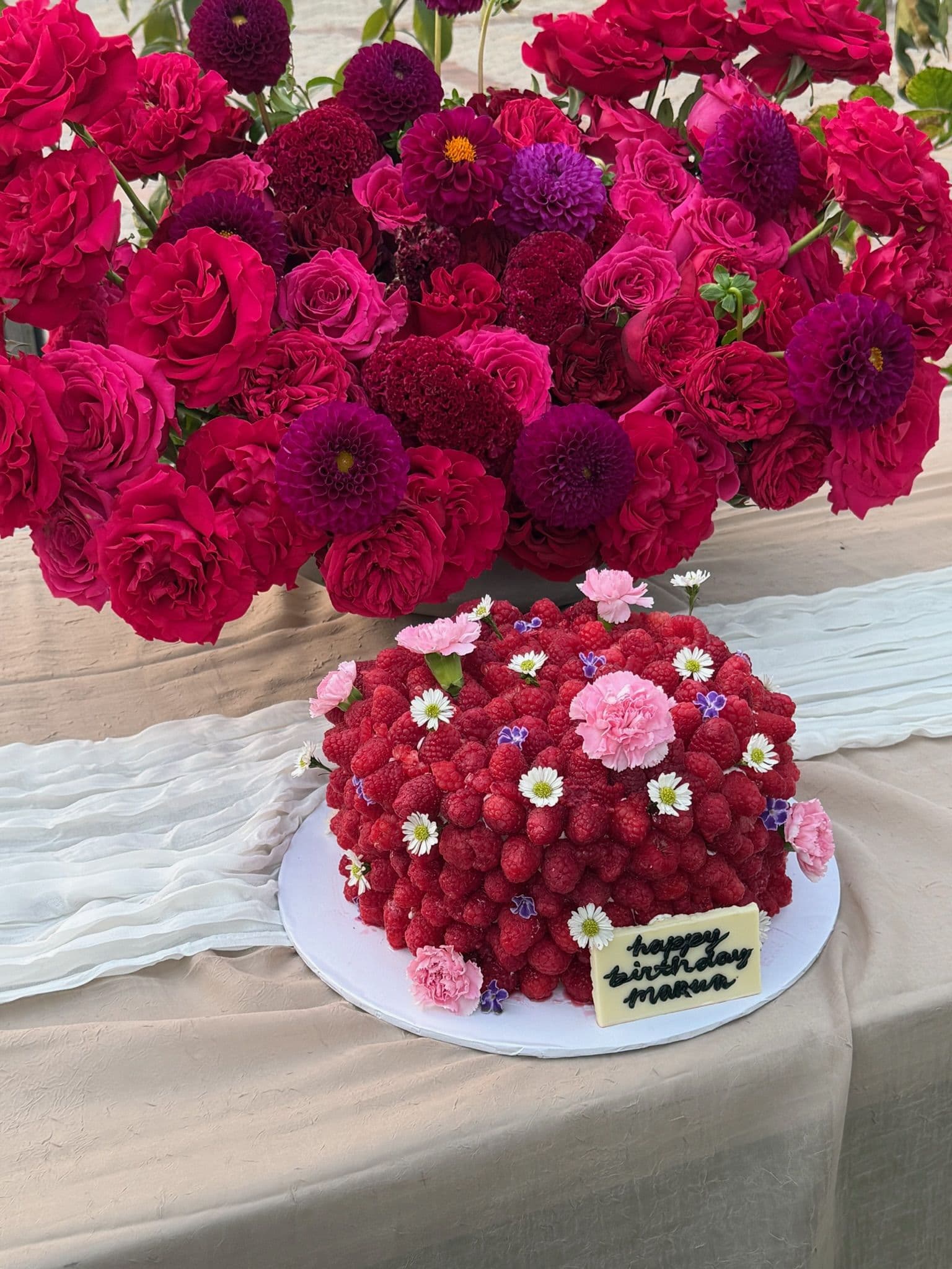 Raspberry-covered cake with a chocolate plaque, framed by crimson and purple dahlias