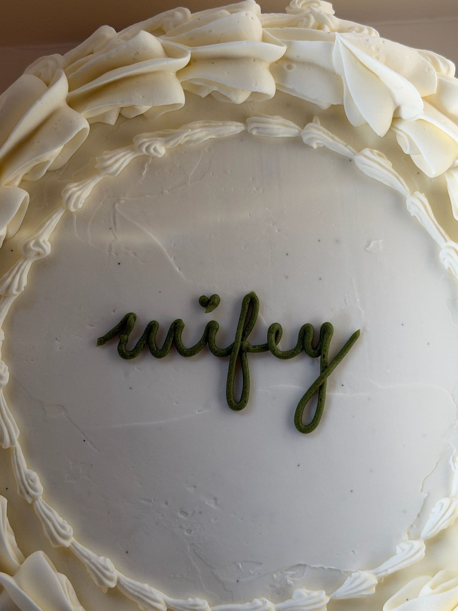 Round cake viewed from above, ruffled buttercream border and "wifey" piped in matcha-green script