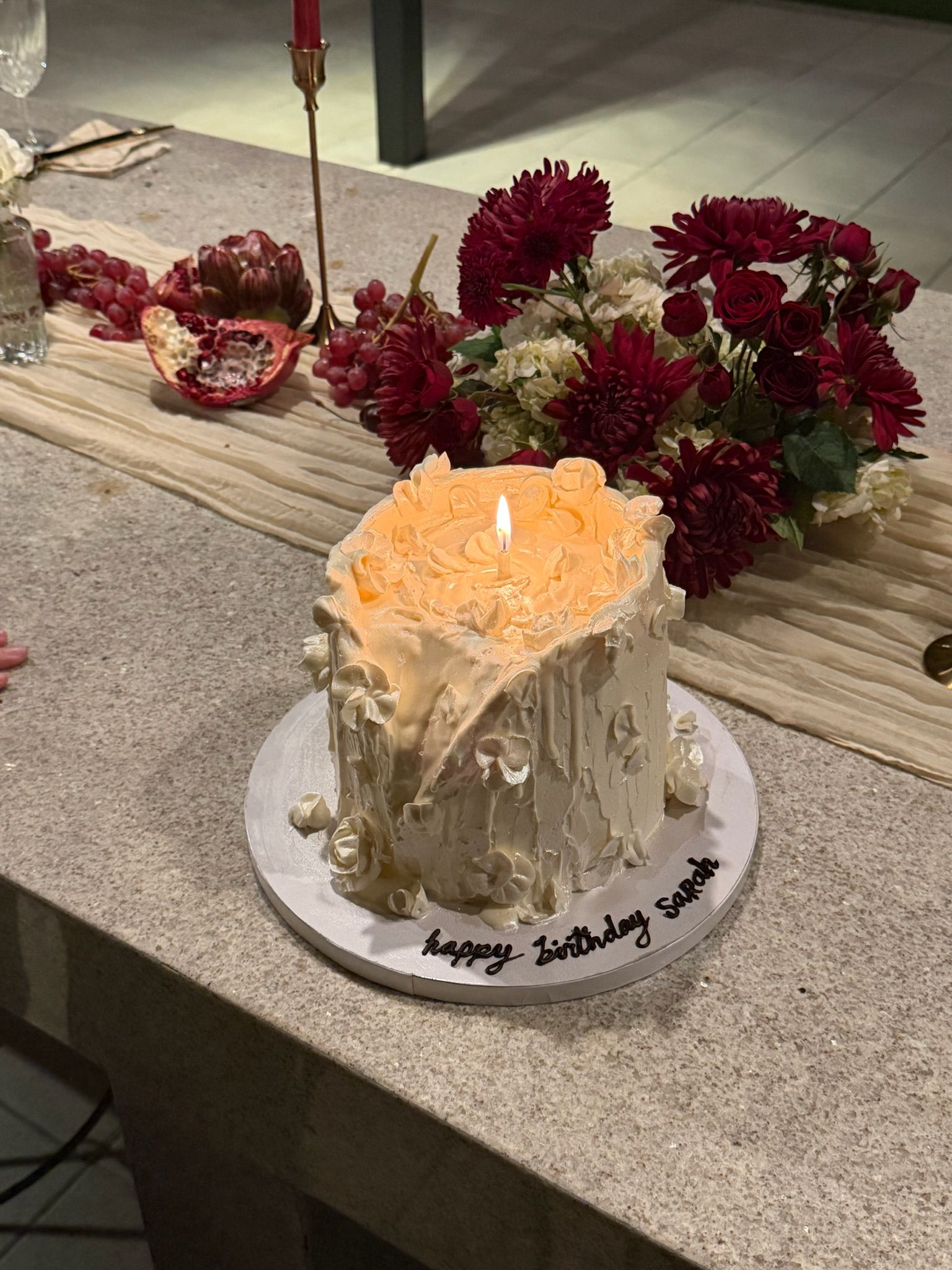 Tall buttercream cake with piped blossoms lit by a single candle, deep red florals behind