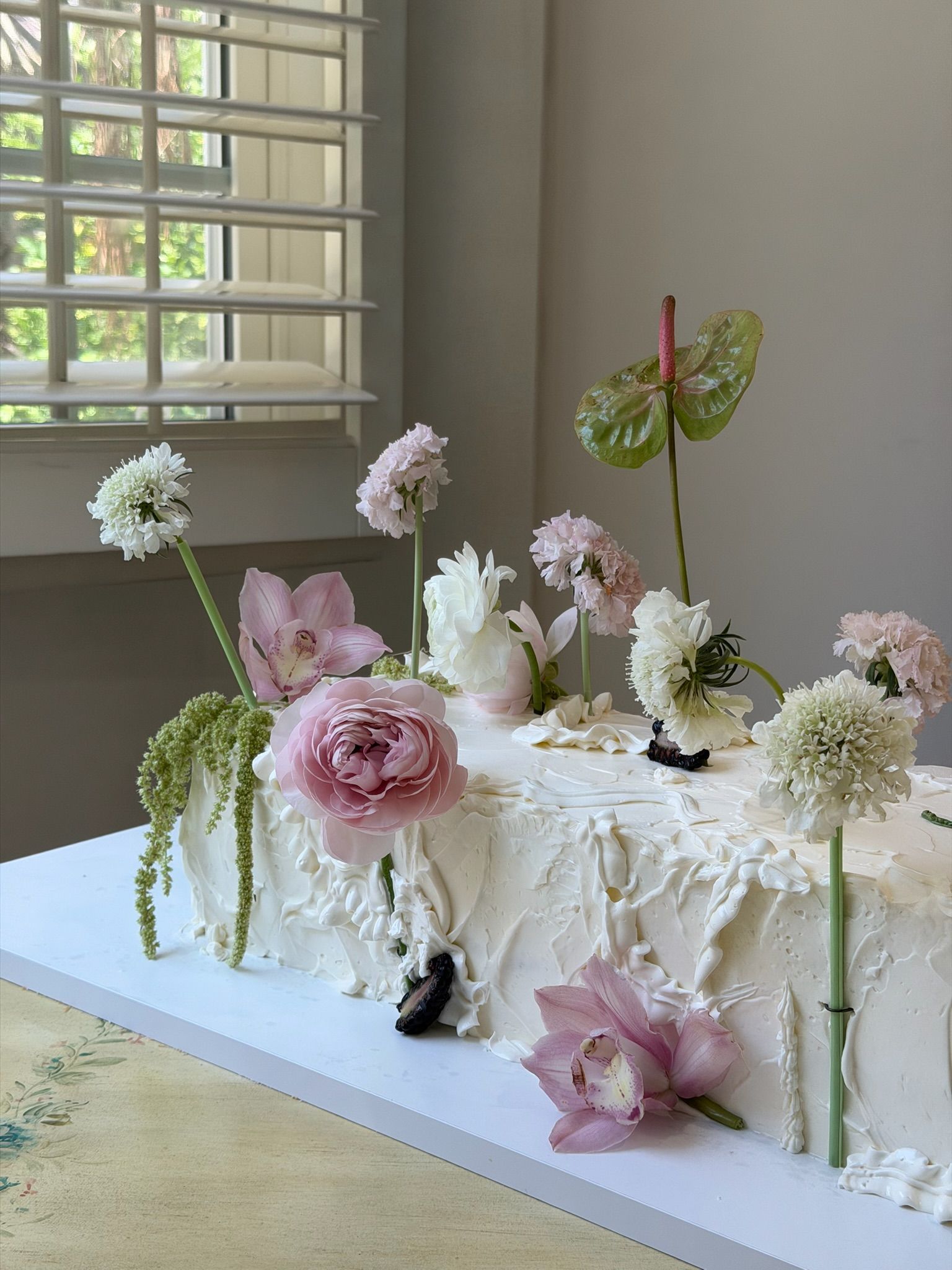 Long ivory sheet cake in textured buttercream, planted with pink orchids, ranunculus and scabiosa on tall stems