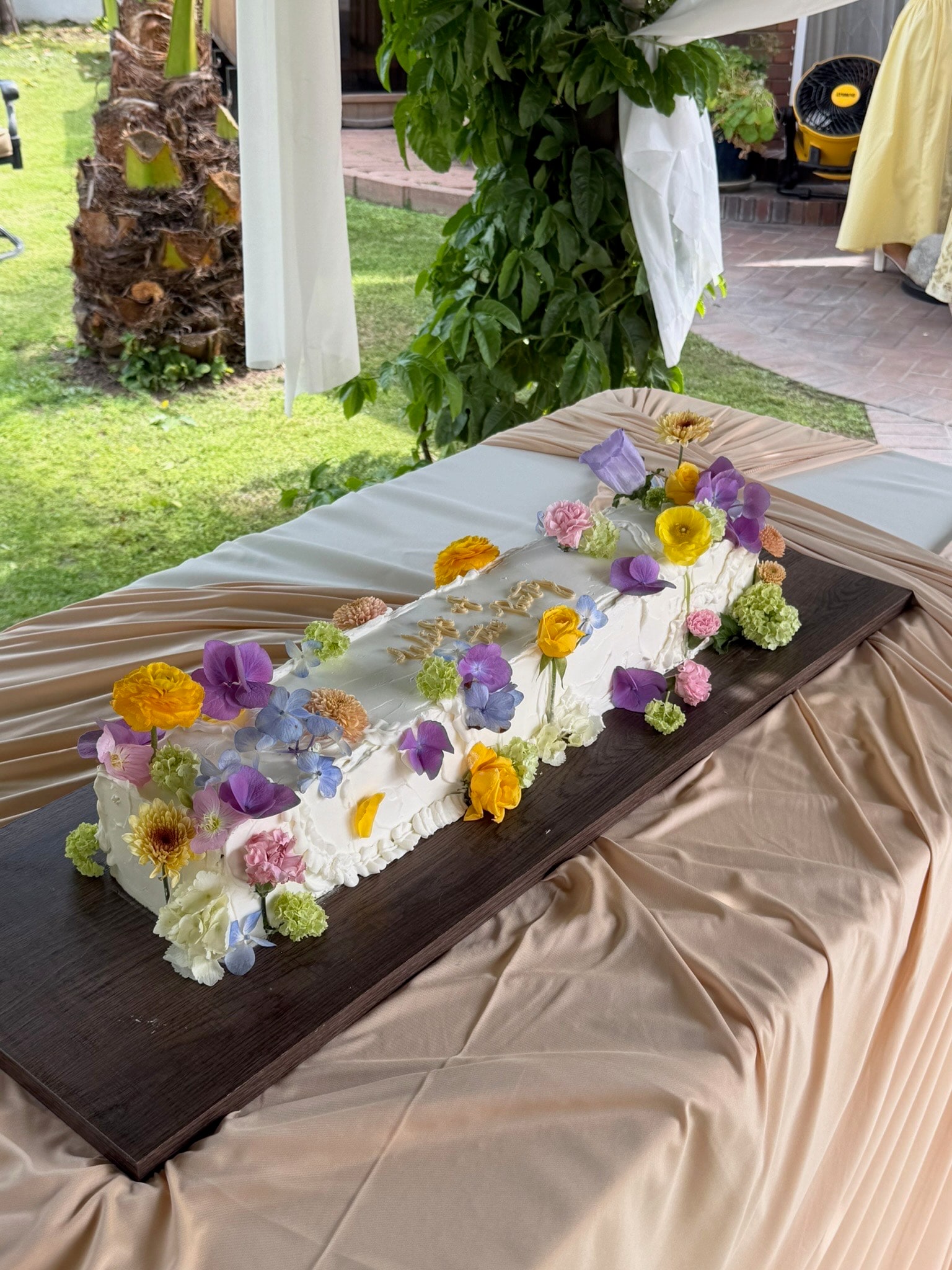 Long sheet cake scattered with purple, yellow and pink garden flowers, on a draped outdoor table