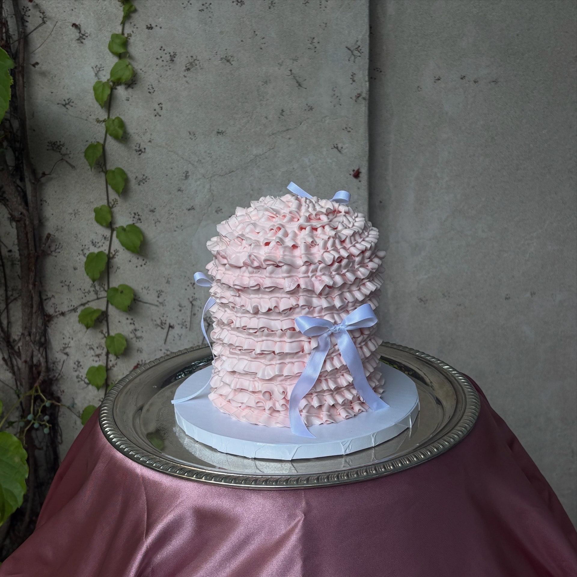 Pink ruffled dome cake trimmed with small white bows, on a silver tray against an ivy wall